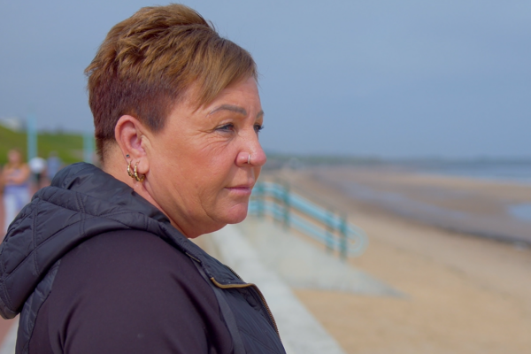 Side profile shot of Claire standing at the coast looking out to sea wearing a black hooded jacked.